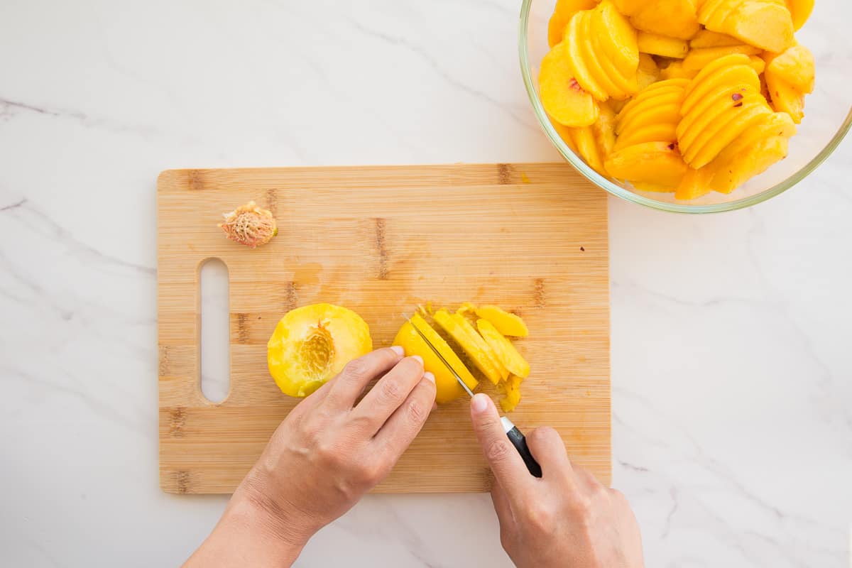 Hands use a knife to slice the fresh peaches on a wooden cutting board to put them in a clear glass mixing bowl in the upper right of the photo.