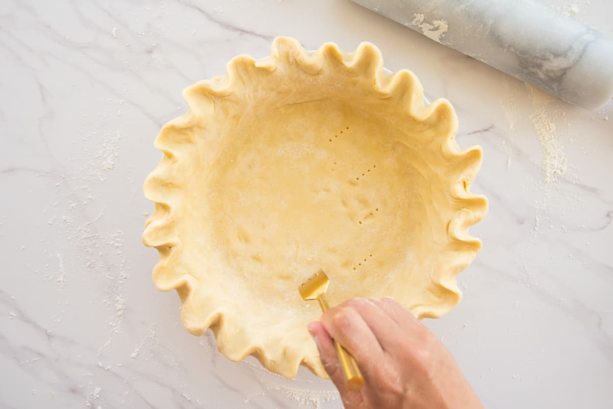 A hand uses a gold fork to pierce holes into the pie dough in a pie plate.