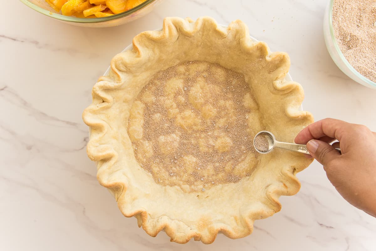 A hand uses a measuring spoon to sprinkle some of the thickening mixture onto the bottom of the par-baked pie shell.