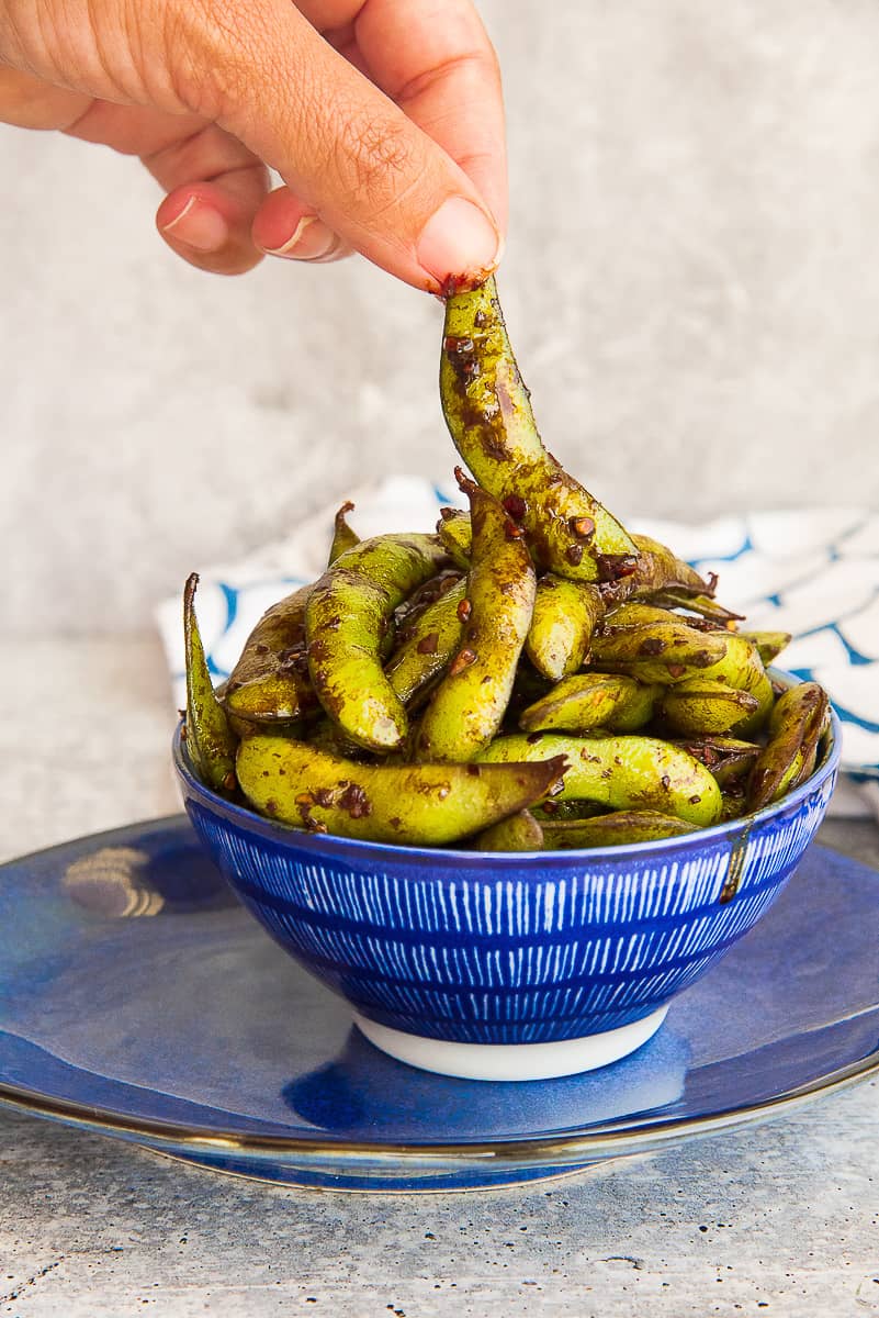 A hand lifts a Spicy Garlic Edamame pod from a blue bowl.