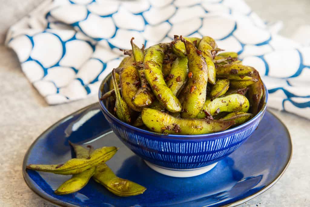 A blue bowl of seasoned edamame sits on a matching blue plate. The edamame is garnished with spices, and a white cloth with blue patterns is seen in the background.
