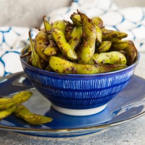 Empty pods are placed on a plate under a blue bowl of Spicy Garlic Edamame.