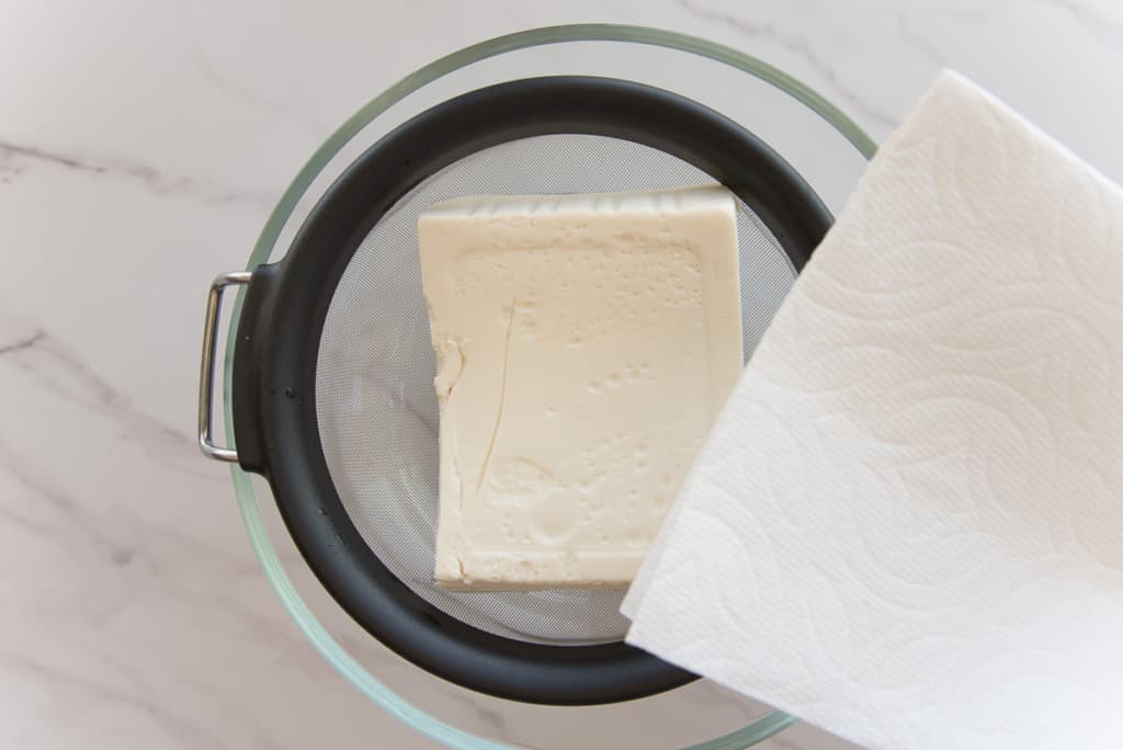 Silken tofu is placed into a fine-mesh sieve to drain before being covered in a paper towel.