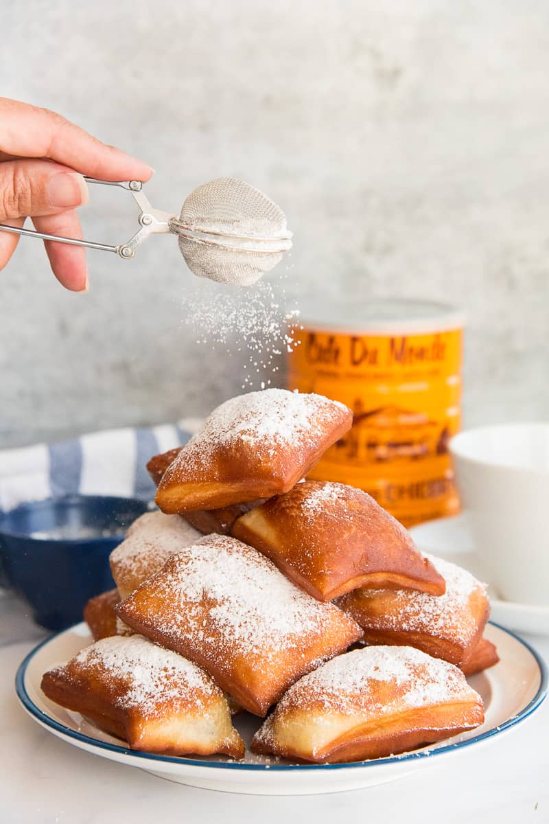 A hand sprinkles powdered sugar over a pile of Beignets on a white blue-rimmed plate.