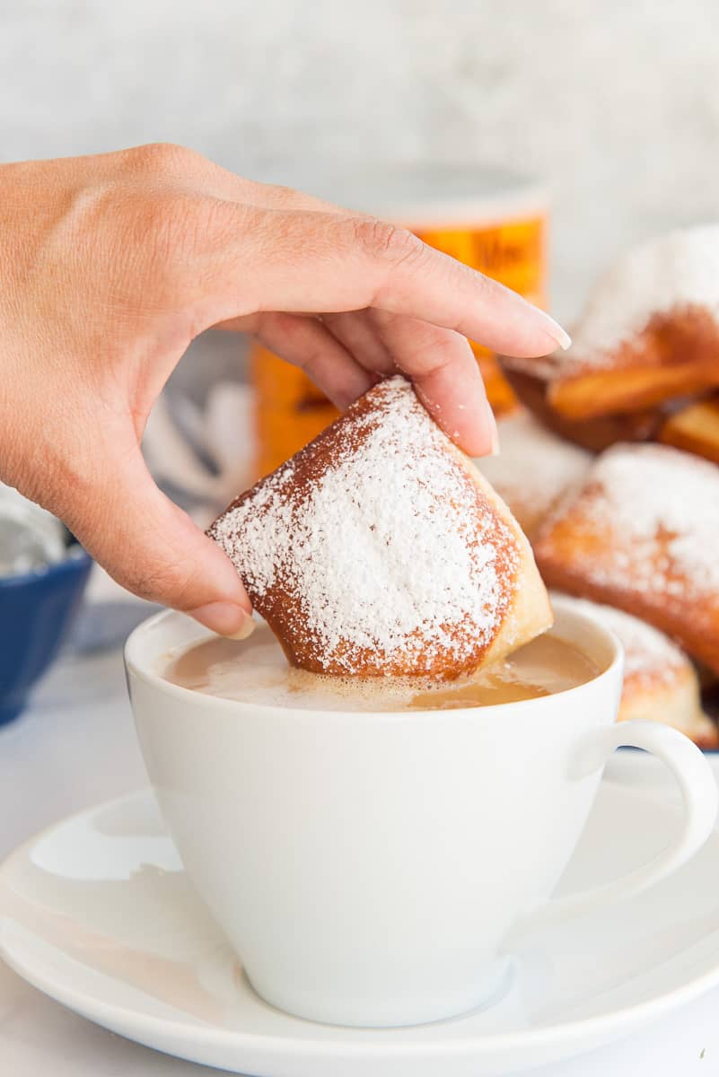 A hand dips a Beignet into a white mug of café au lait.
