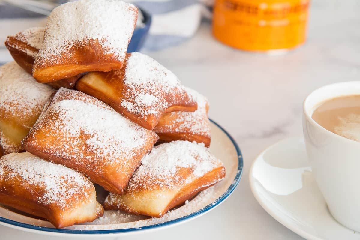 A pile of sugary Beignets are stacked on a white, blue-rimmed plate next to a mug of café au lait.