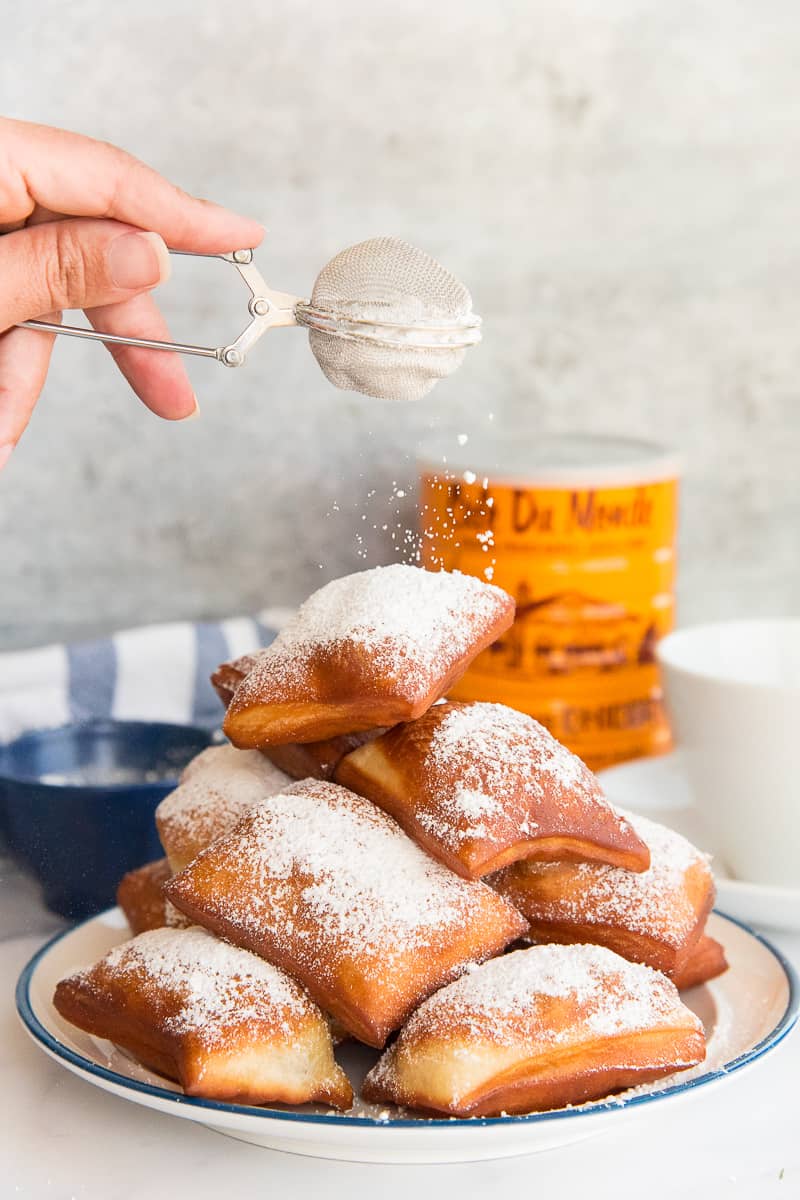 A hand sifts powdered sugar from a tea strainer over a pile of Beignets.