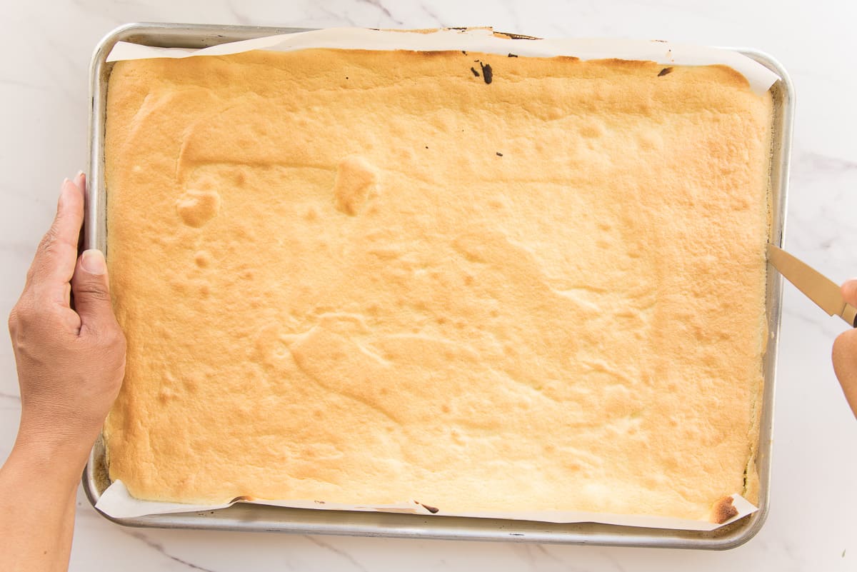 A hand uses a paring knife to loosen the cake from the sides of a sheet pan.