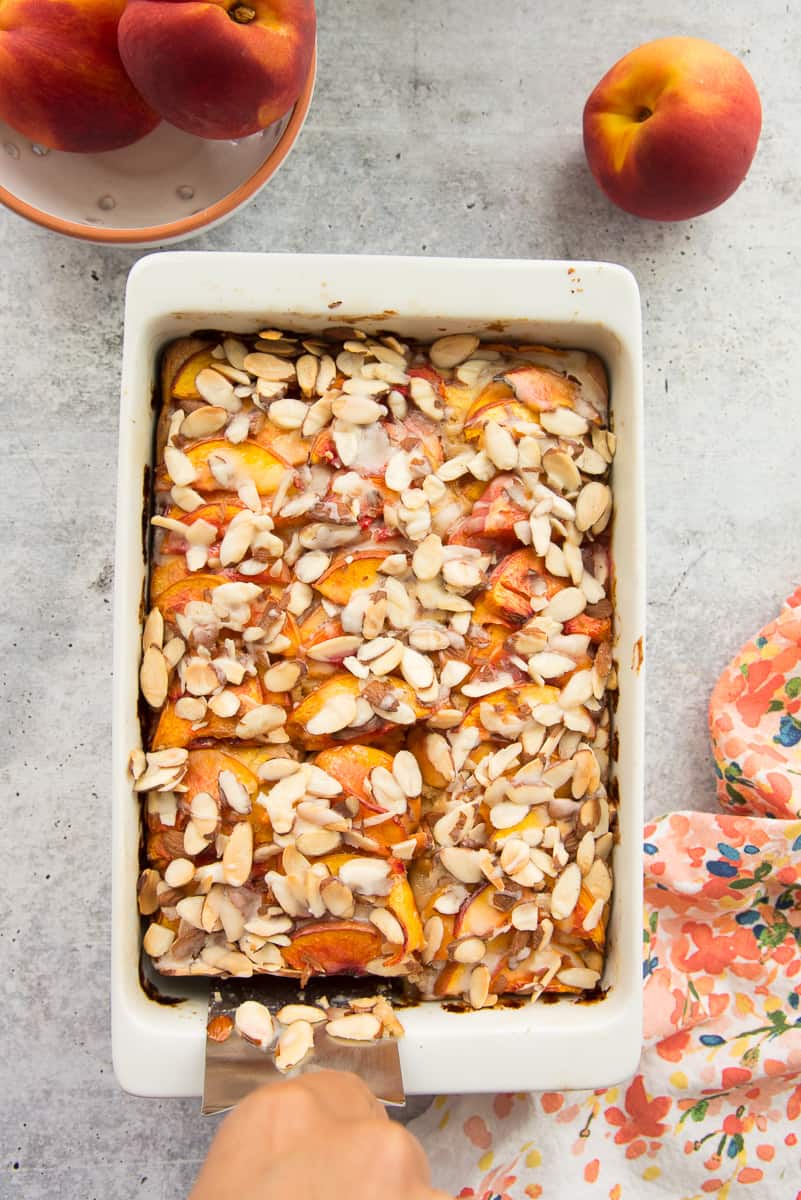 A hand slides a silver spatula under a piece of the Nectarine Almond Coffee Cake in a white baking dish.