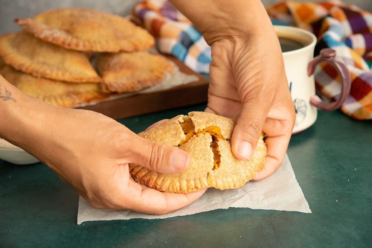 Hands break apart a Pumpkin Cheesecake Empanadas to reveal the flaky texture of the exterior and the creamy interior.