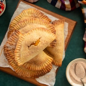 A stack of Pumpkin Cheesecake Empanadas on a wooden board next to a bowl of spiced sugar.
