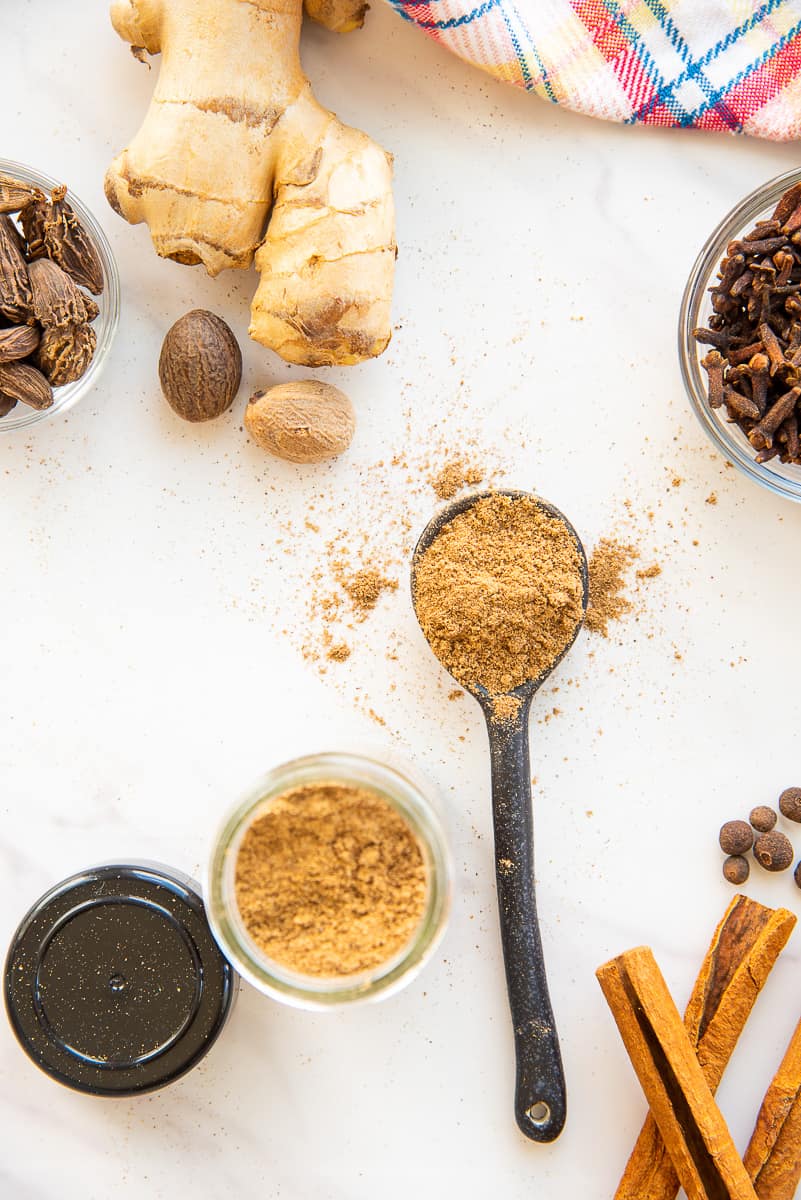 The Chai Spice Blend on a black spoon surrounded by the ingredients to make the blend.