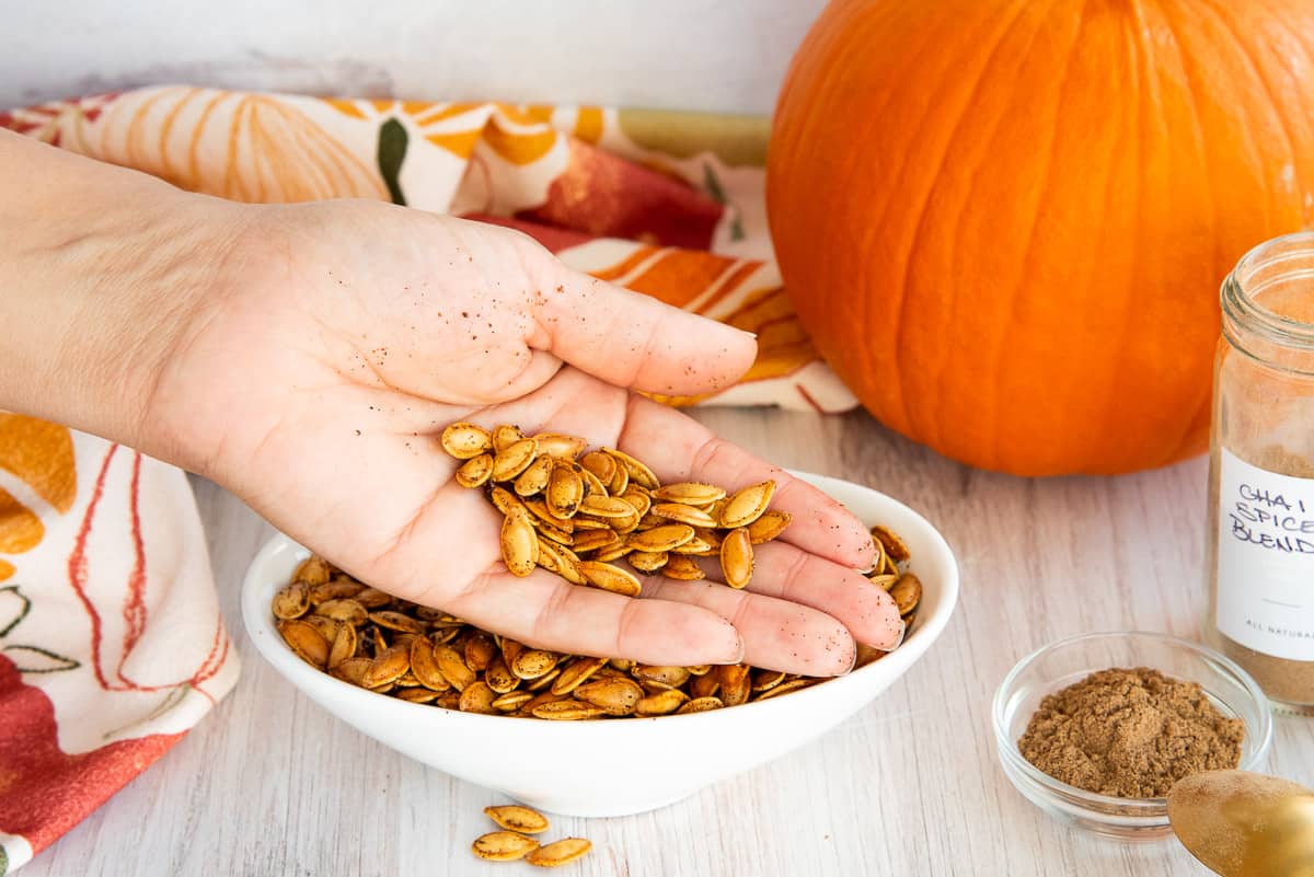 A hand lifts Chai Spiced Pumpkin Seeds from a white bowl.