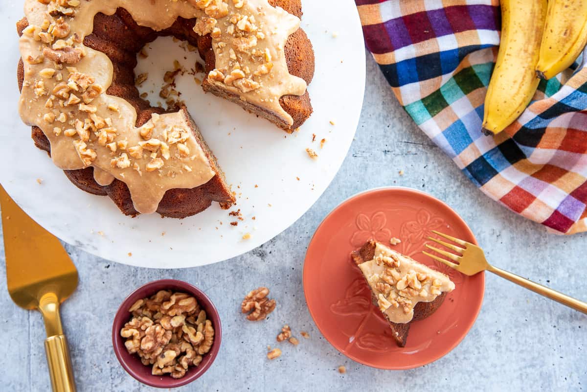 A slice of Maple Walnut Banana Bundt Cake on an orange plate next to a cake stand with the rest of the cake.