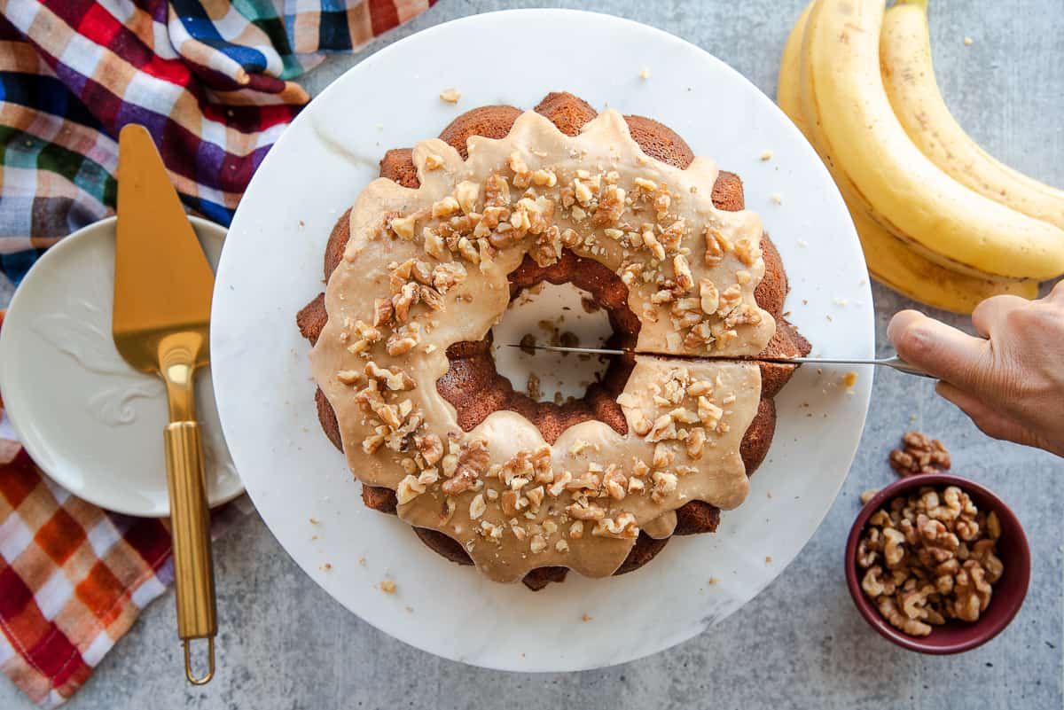 A hand uses a knife to cut a slice of the Maple Walnut Banana Bundt Cake from the cake.