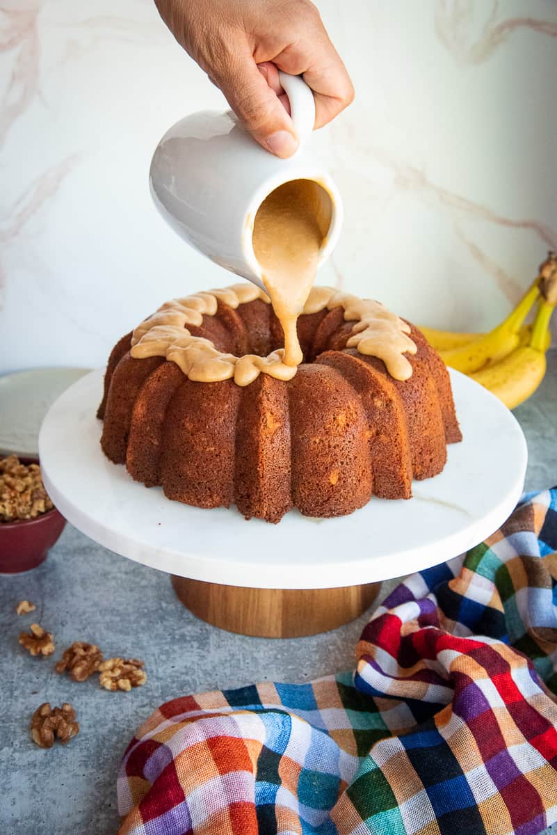 A hand pours the Brown Butter Caramel Glaze from a white pitched onto the Maple Walnut Banana Bundt Cake