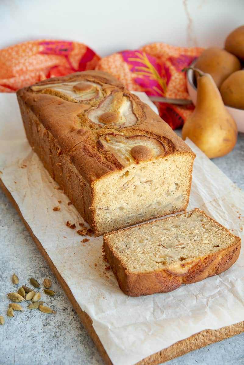 A loaf of Pear Cardamom Bread with one slice laying down in front of it on a wooden board.