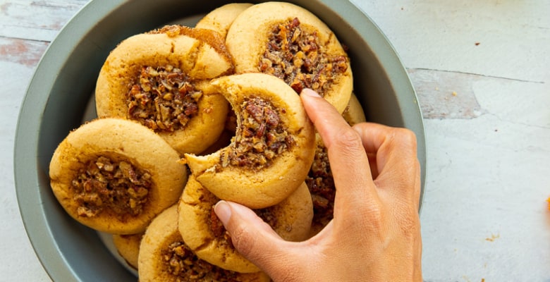 A hand lays a bitten cookie on top of a pile of Pecan Pie Thumbprint Cookies in a metal pie tin.