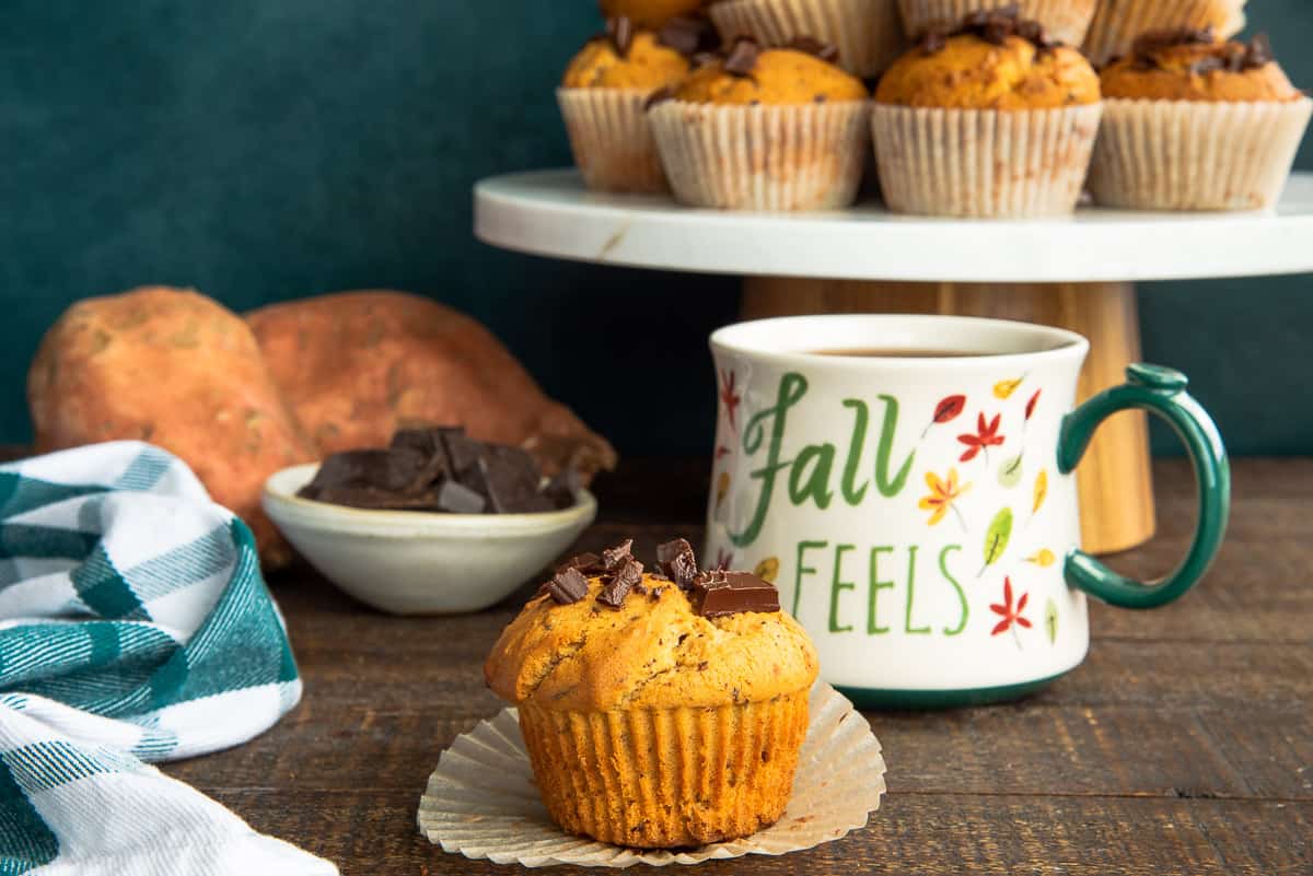 A muffins sits in front of a mug of coffee and a pile of Sweet Potato Chocolate Chunk Muffins.