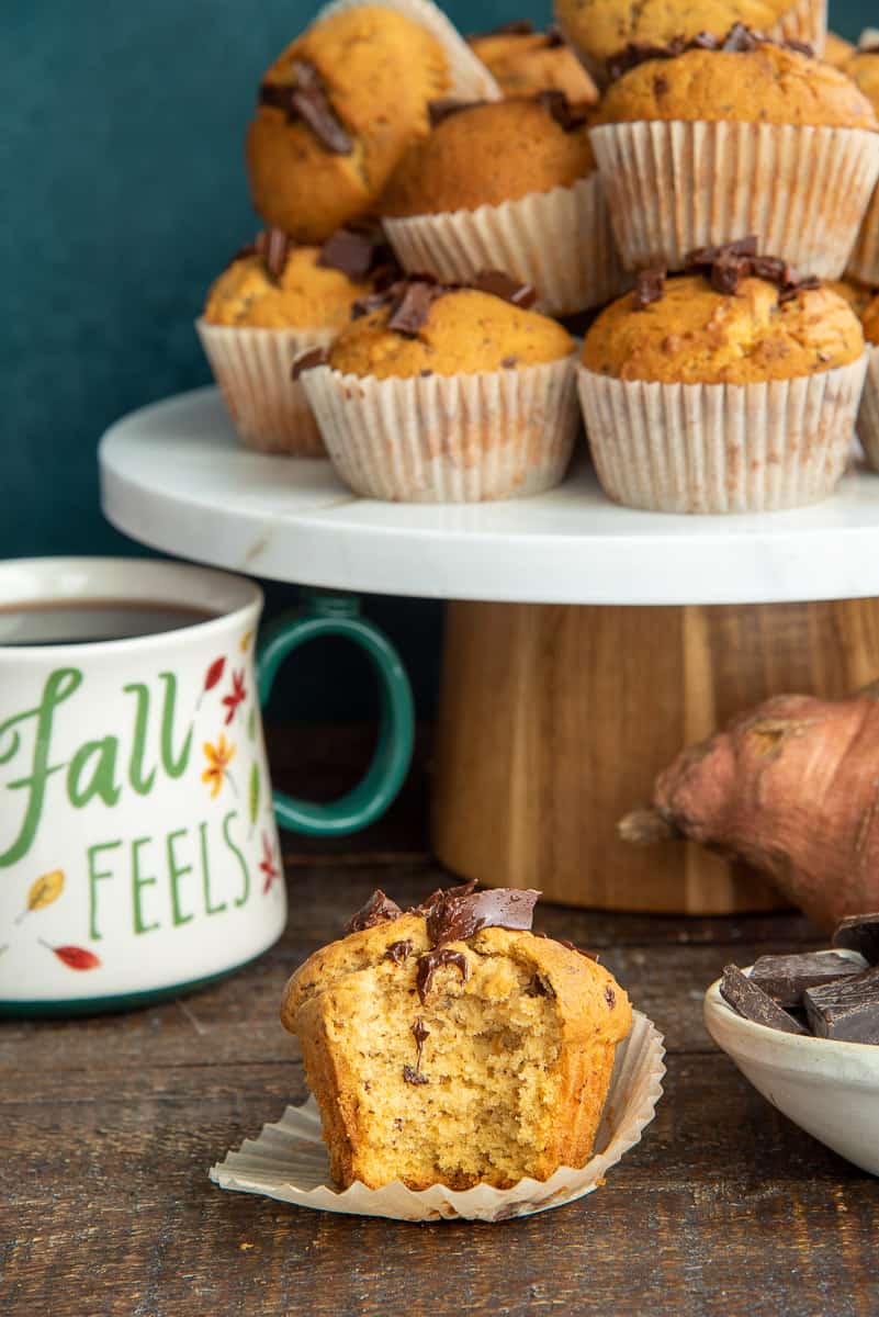 A single Sweet Potato Chocolate Chunk Muffin with a bite taken out is in front of a cake stand piled with Sweet Potato Chocolate Chunk Muffins.