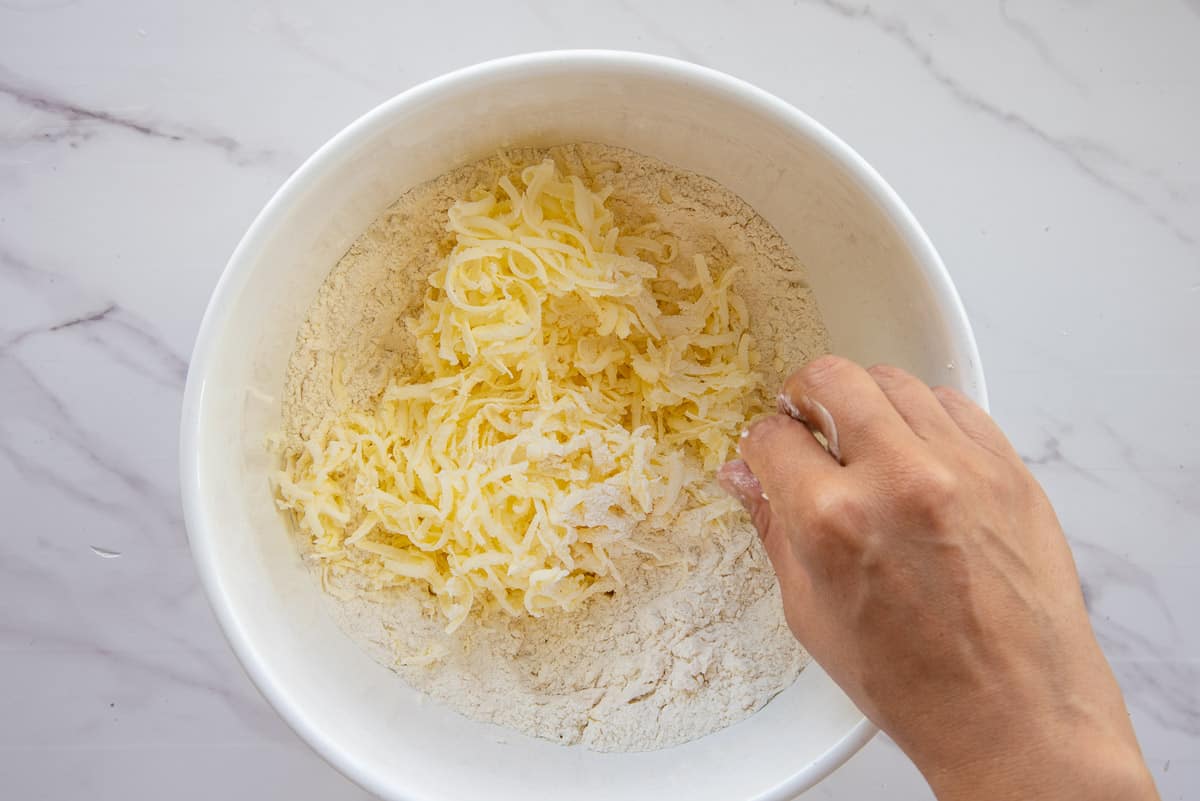 Shredded butter is cut into the dry ingredients mixture in a white ceramic bowl.