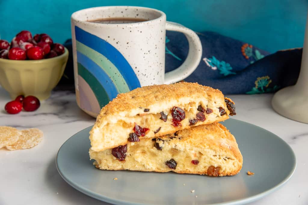 A plate with two cranberry scones stacked on top, set next to a mug of coffee. In the background, there's a bowl of cranberries and a dark cloth with a floral pattern. The scones have a golden crust and visible cranberry pieces.