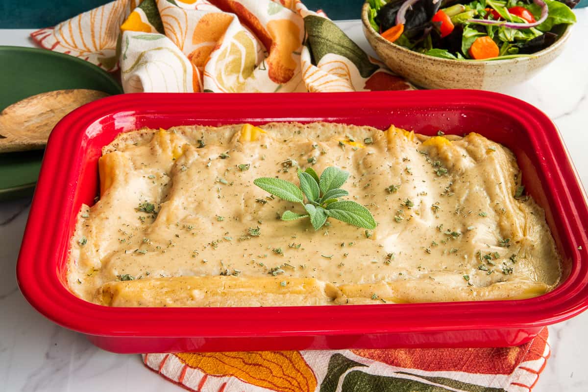 The baked Pumpkin Manicotti in Béchamel Sauce in a red casserole is garnished with fresh sage.
