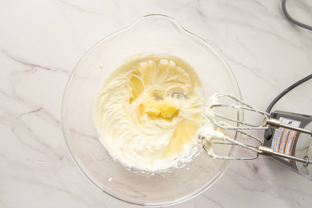 The butter is added to the cream cheese mixture in a clear glass mixing bowl.