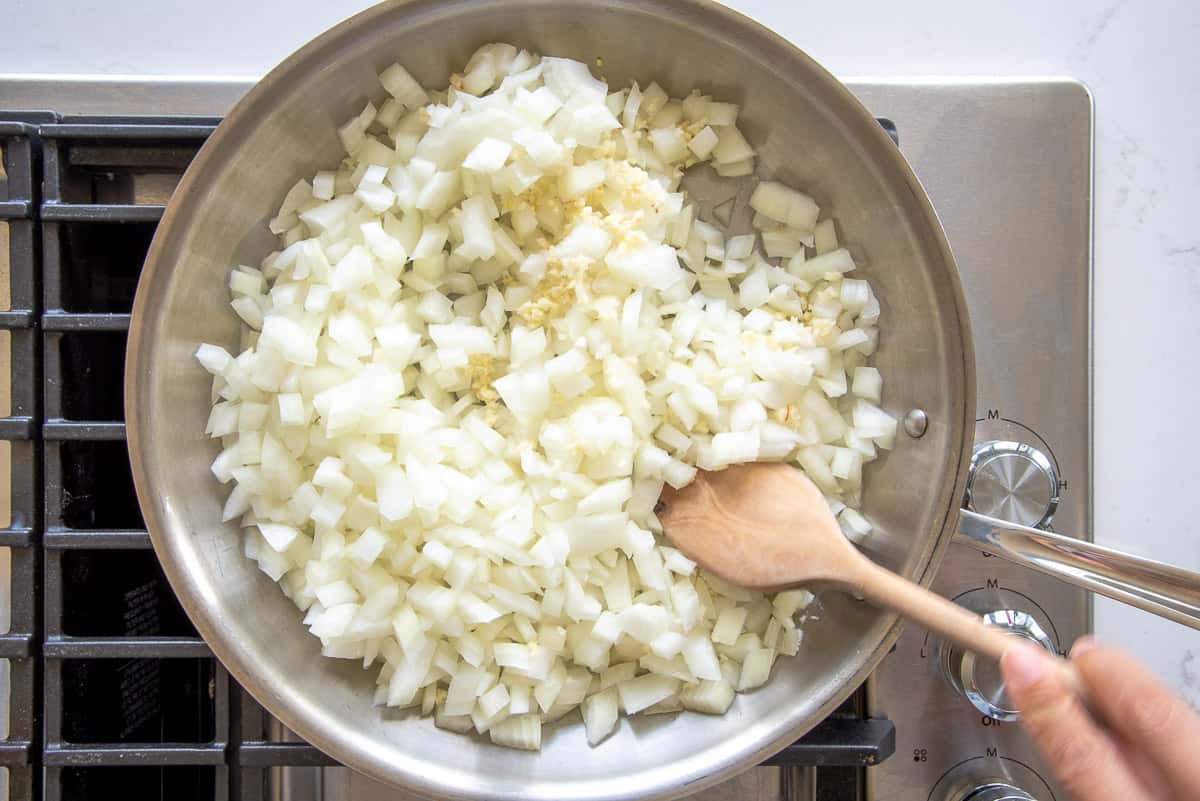 Garlic and diced white onions are stirred together in a silver sauté pan.