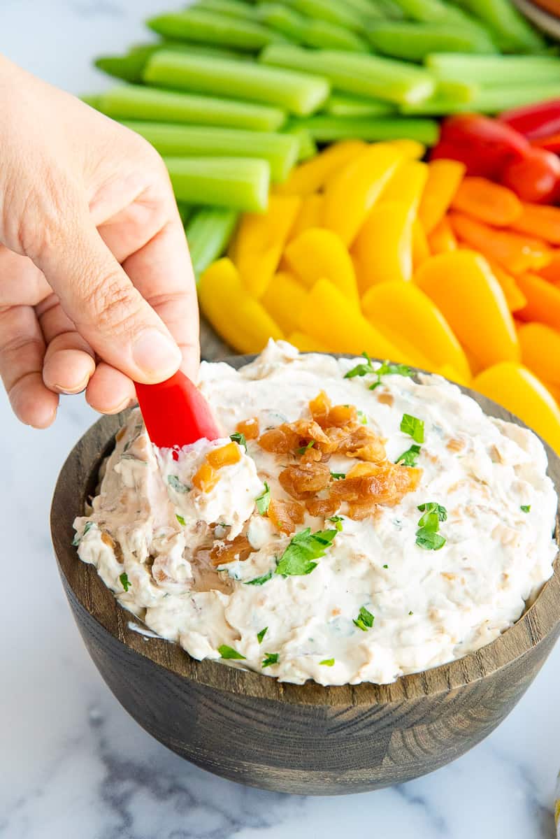 A hand dips a red snacking pepper slice into a bowl of Caramelized French Onion Dip .