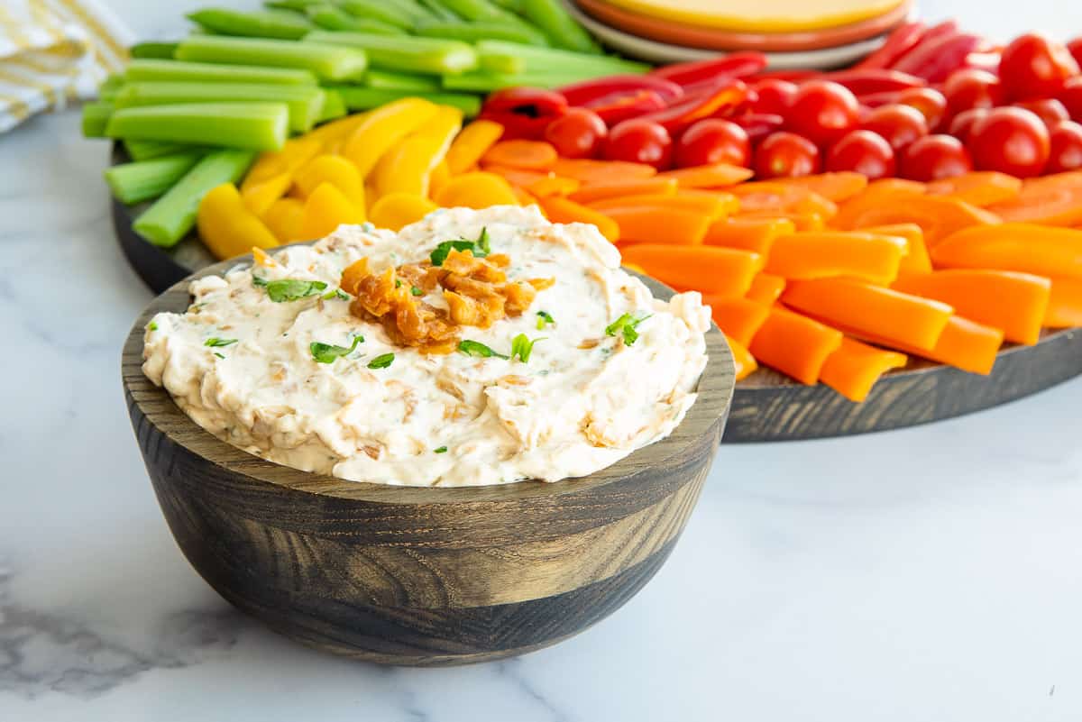 A bowl of Caramelized French Onion Dip in front of a wooden platter of crudité.