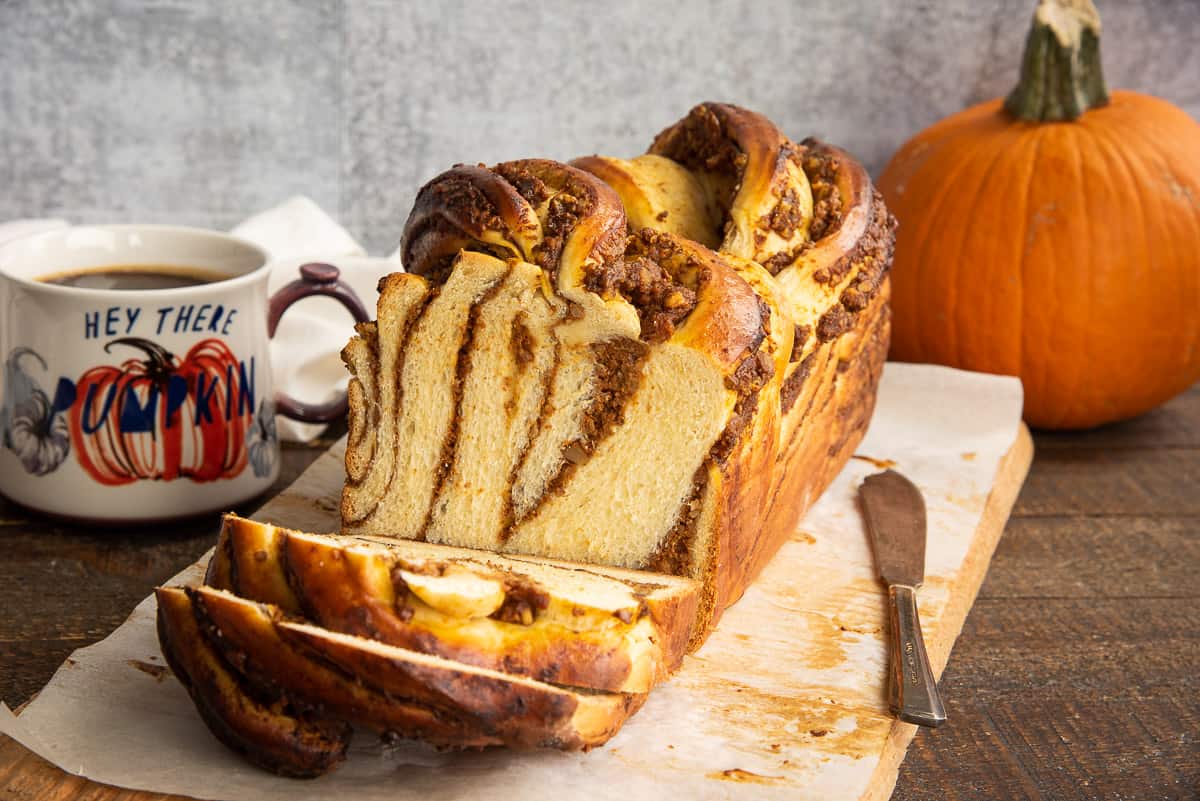 The loaf of Pumpkin Pecan Babka is sliced in front of a whole pumpkin and a mug of coffee.