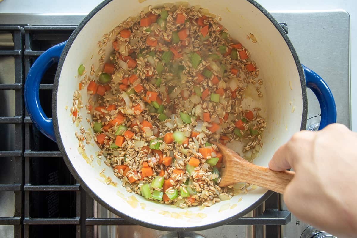 The farro is stirred into the mirepoix with a wooden spoon in a dutch oven.