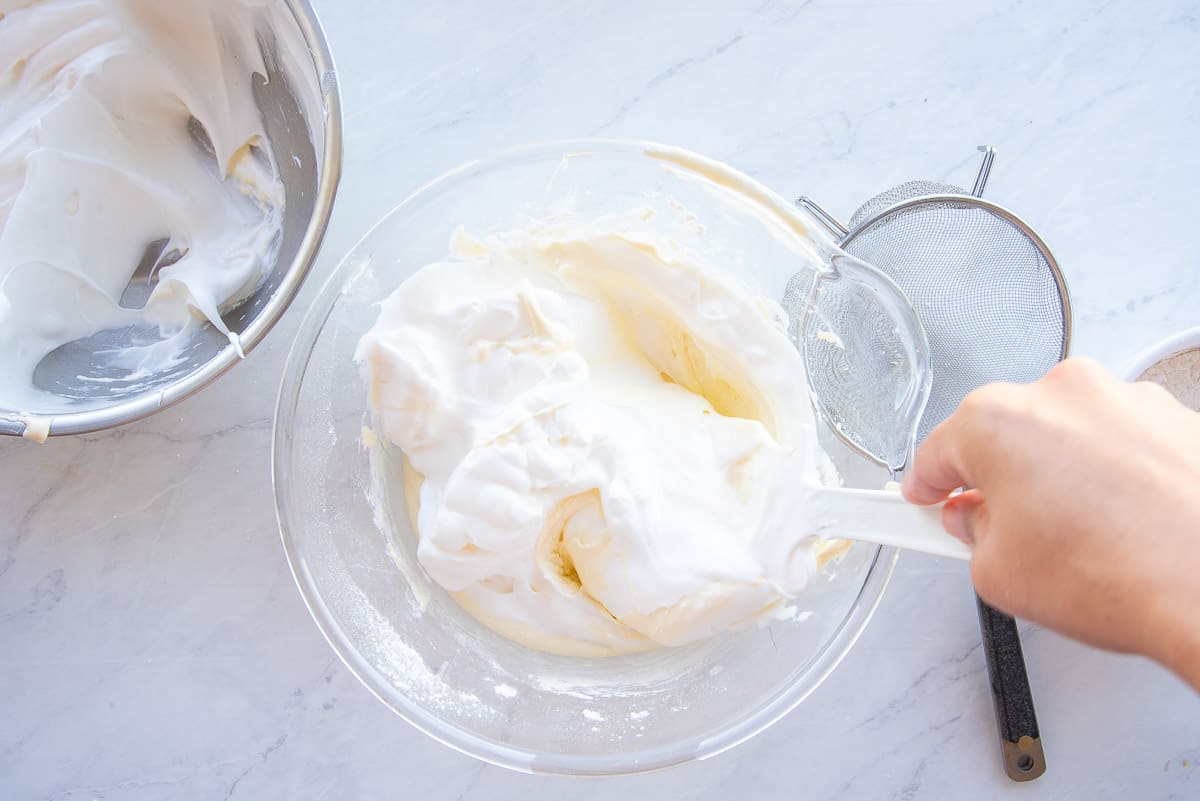 The egg whites and flour are gently folded into the whipped egg yolks in a clear glass bowl.