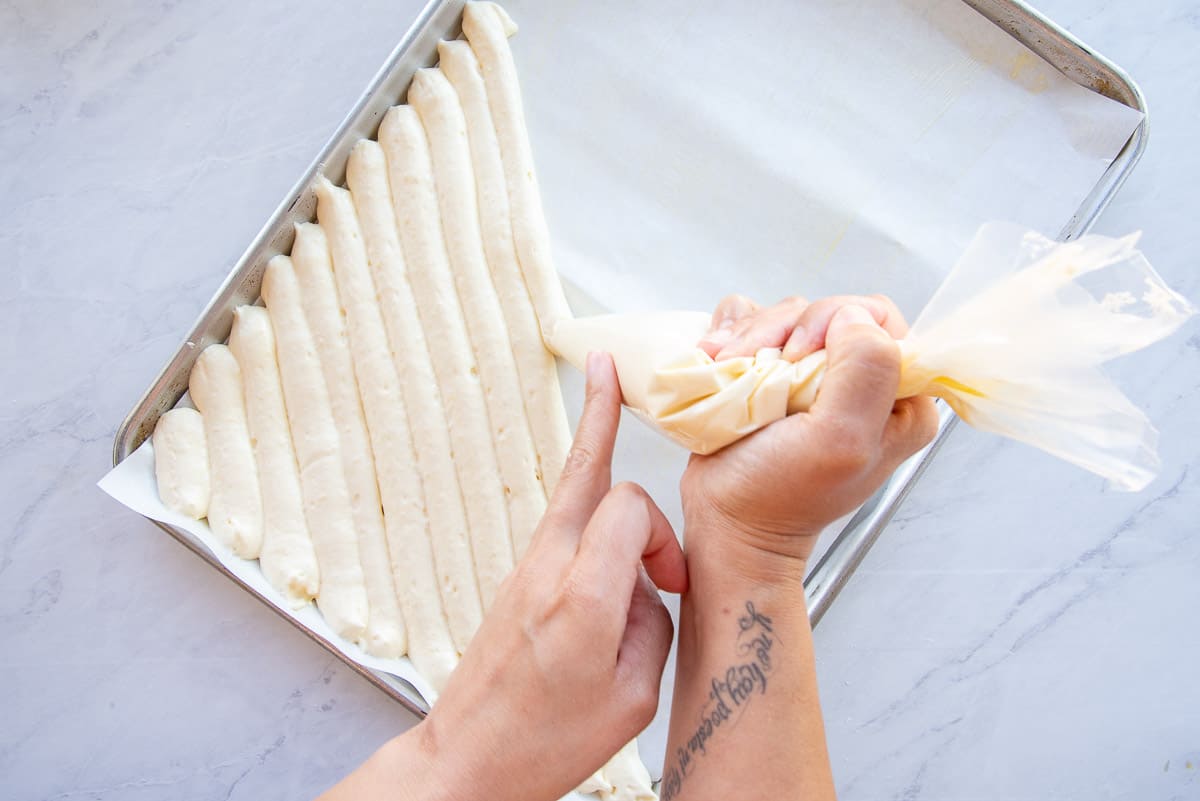 Hands hold a plastic piping bag to pipe the ladyfinger sponge batter onto a parchment-lined sheetpan.