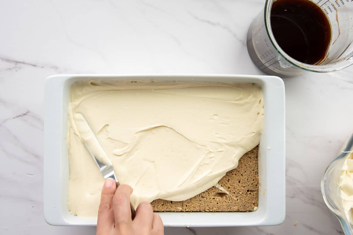 A hand uses a small offset spatula to spread the mascarpone filling over the espresso-soaked sponge layer.