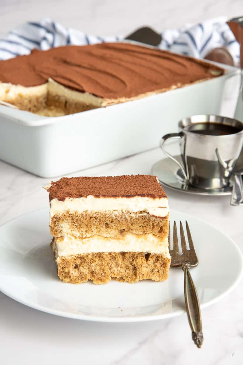 A white plate with a serving of Classic Tiramisu from Scratch is in front of a dish of tiramisu and a small silver cup of espresso.