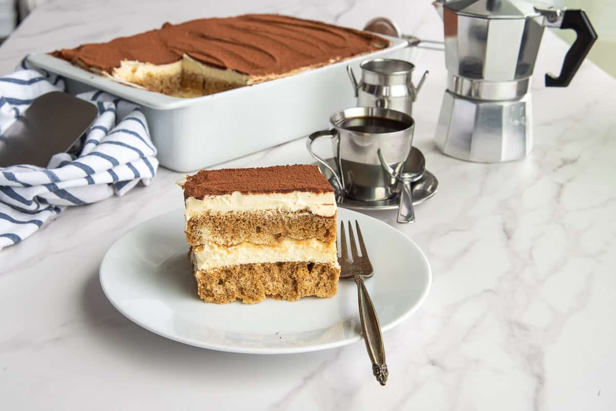 A dish of Classic Tiramisu from Scratch in background. A white plate with a serving of tiramisu in the foreground.