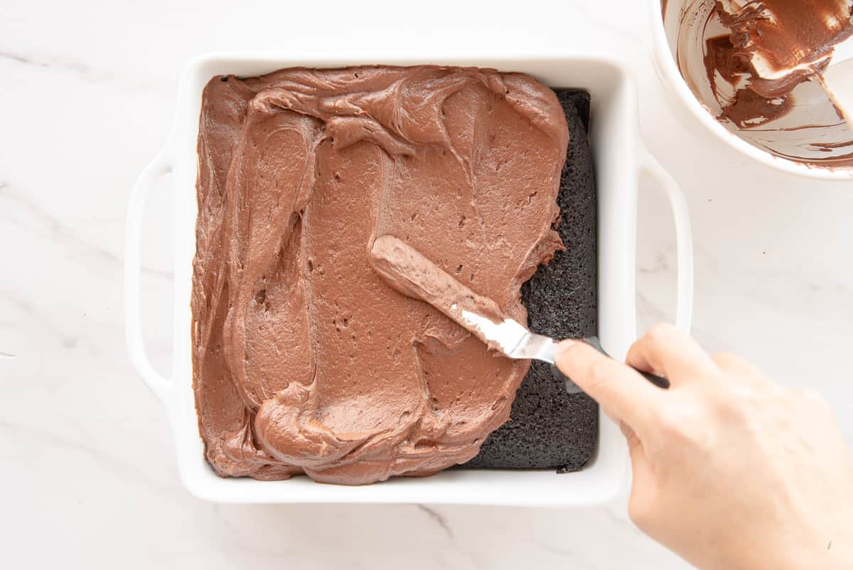 A hand uses an offset spatula to spread the chocolate ganache on the baked dessert.