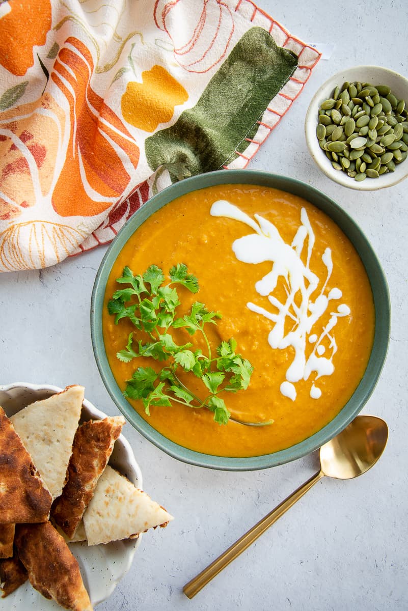 Green bowl of Vegan Curry Pumpkin Soup garnished with cilantro and coconut cream is next to a bowl of pepitas and triangles of naan.