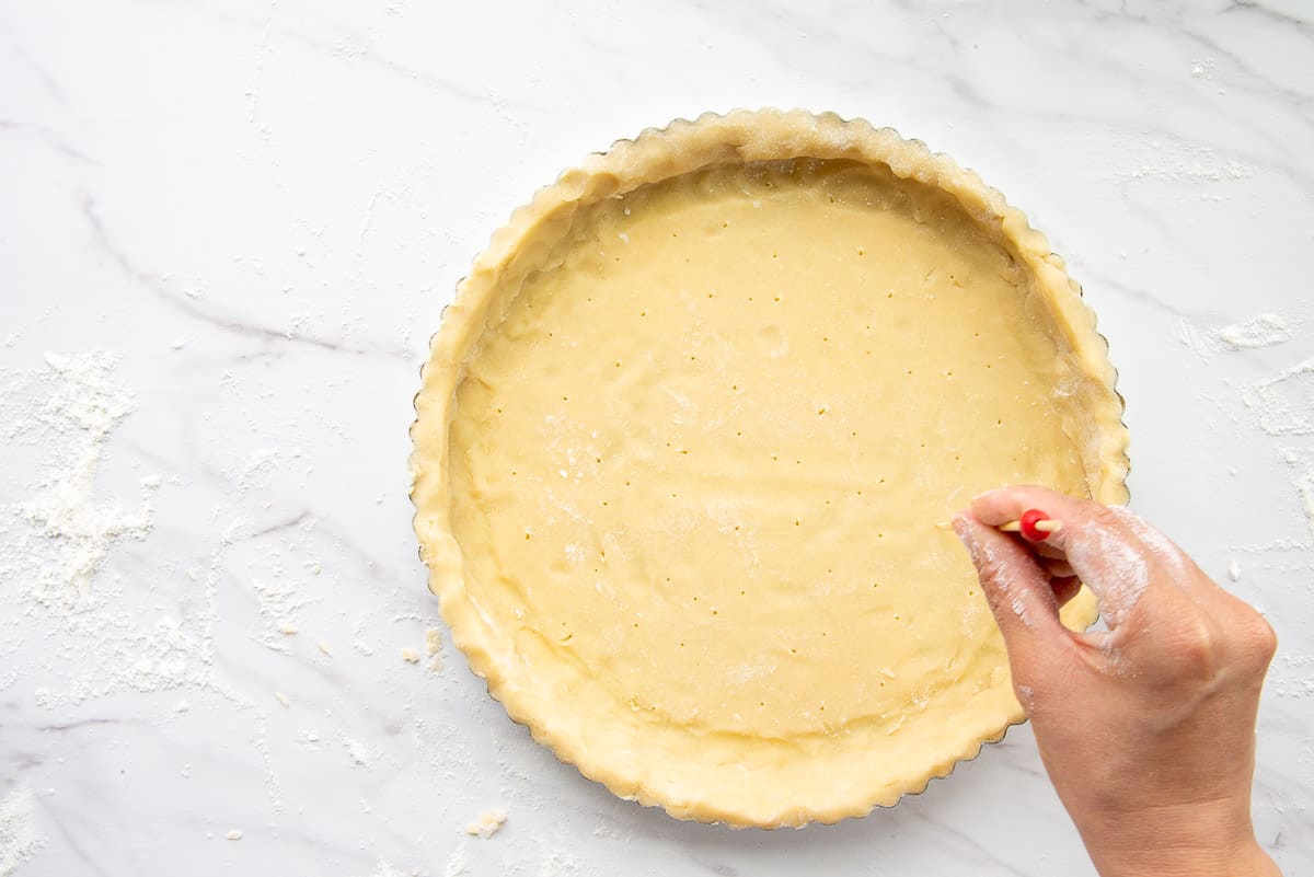 A hand uses a skewer to poke venting holes in the almond tart dough inside a tart pan.