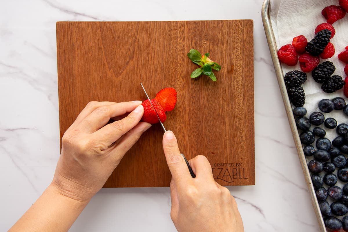 Hands hold and slice a fresh strawberry on a wooden cutting board.