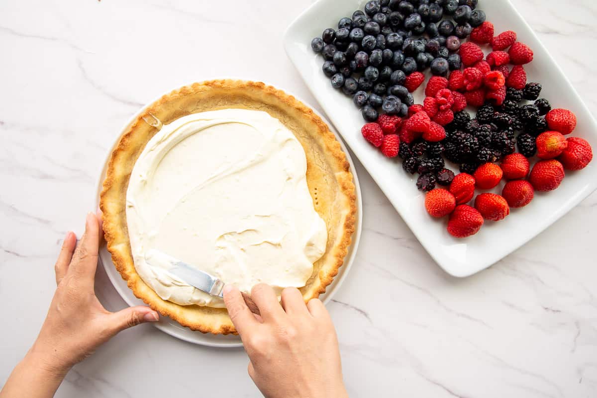 Hands use an offset spatula to spread the mascarpone filling inside of the baked almond tart shell. A platter of fresh berries is on the right side.