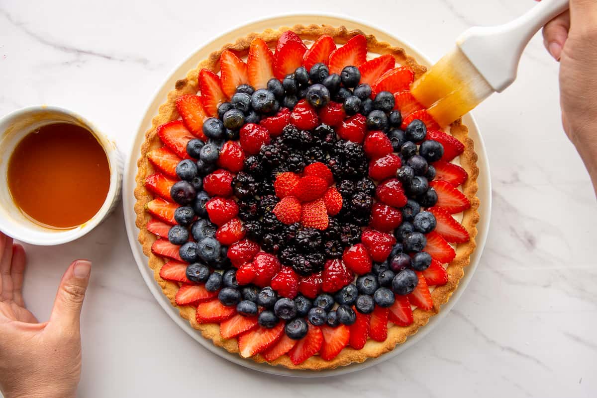 A hand uses a white pastry brush to brush the berries with a fruit glaze.