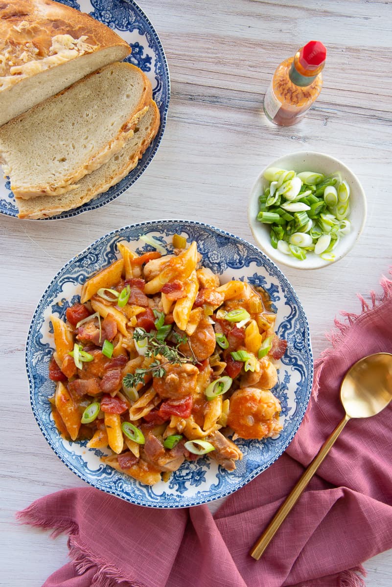 Jambalaya Pasta in a blue and white bowl. A plate of sliced bread is a top left and a mauve napkin is at bottom right.