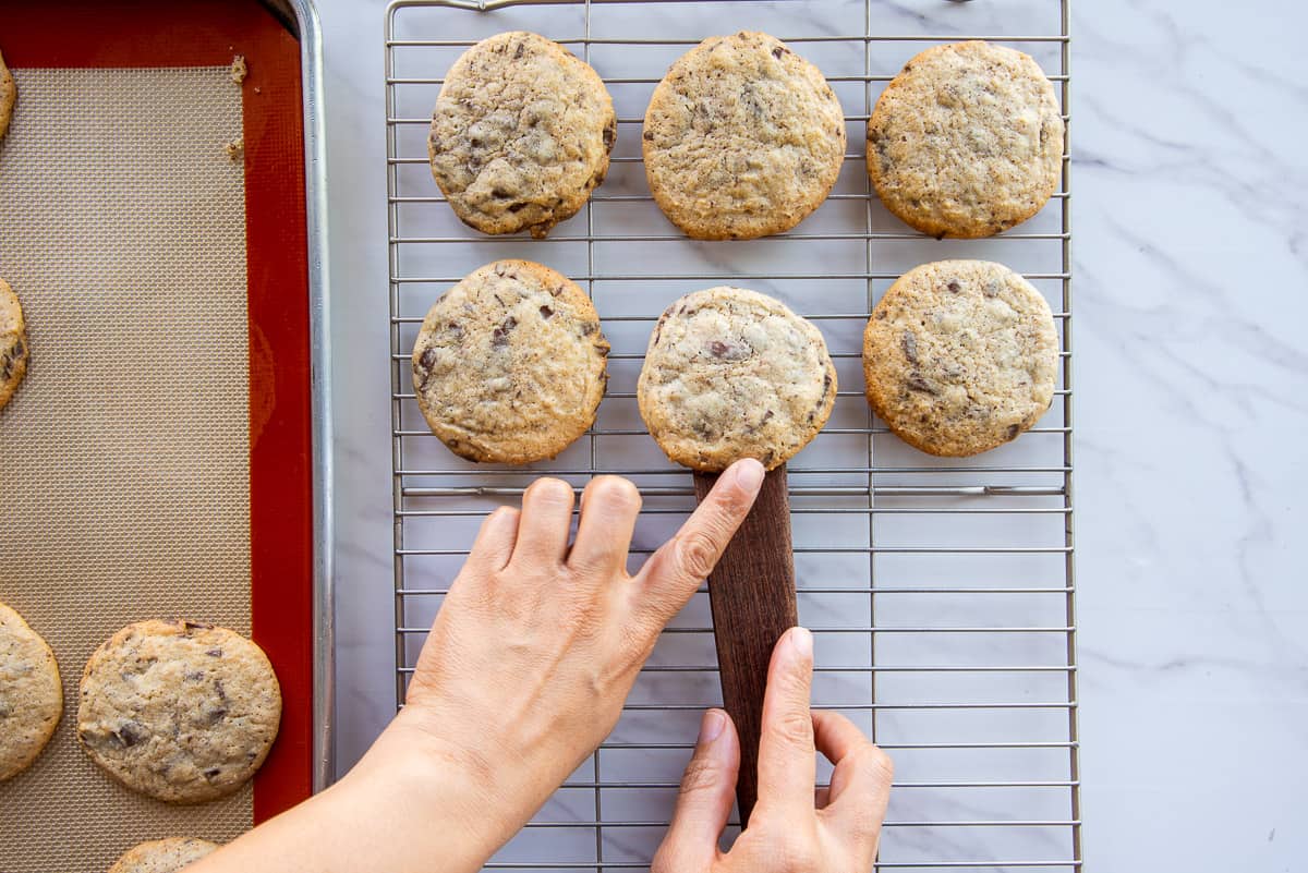 Hands use a flat wooden spatula to transfer the baked cookies from the baking sheet to a cooling rack.