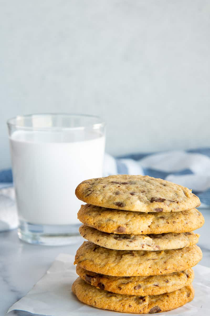 Rye Sourdough Discard Chocolate Chip Cookies stacked on top of each other in front of a glass of milk and a blue and white kitchen towel.
