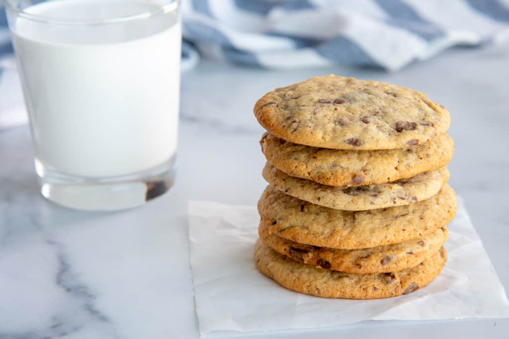 A stack of six chocolate chip cookies is placed on a small white piece of parchment paper on a marble surface. Behind the cookies, there is a glass of milk and a blue-and-white striped cloth.