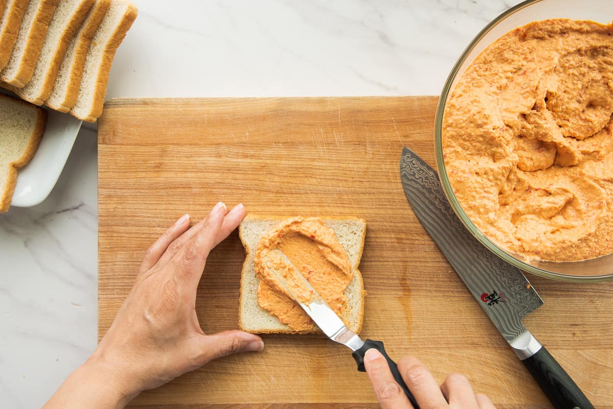One hand holds a piece of bread while the other spreads the mezcla on it with an offset spatula.