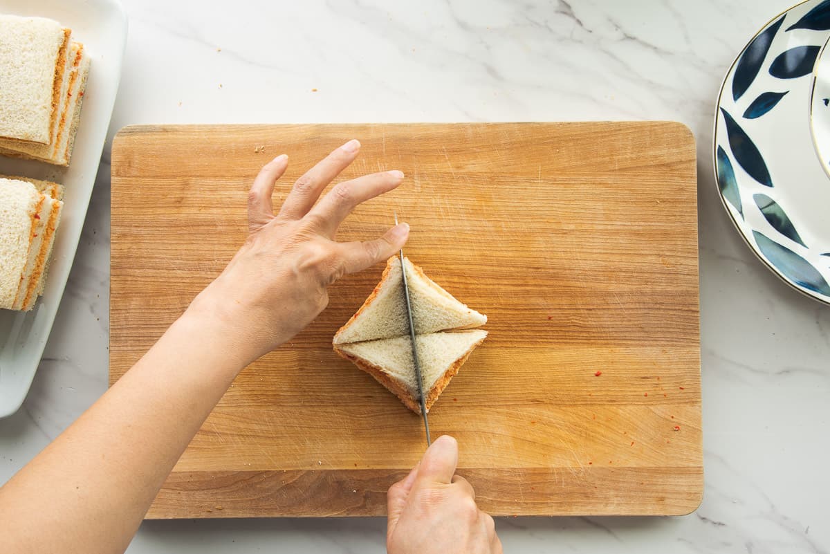Hands cut the whole sandwich into four triangles on a wooden cutting board.